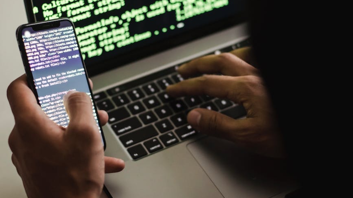 unrecognizable hacker with smartphone typing on laptop at desk